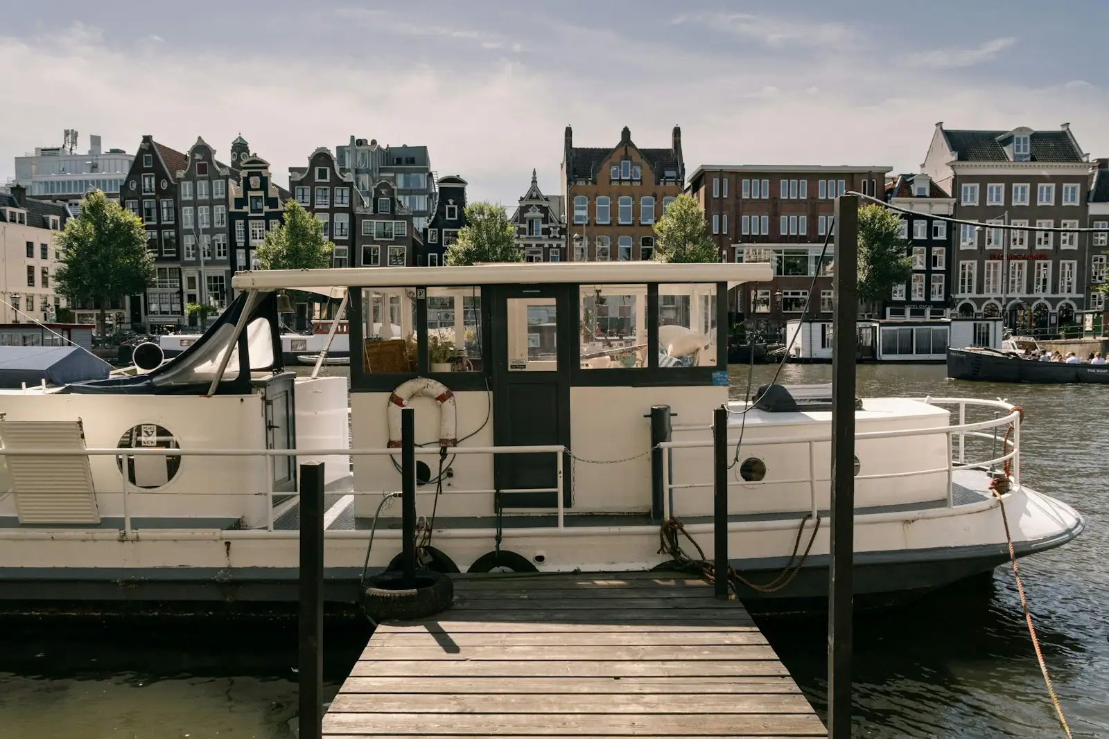 A white houseboat moored at a wooden dock in Amsterdam with historic canal houses