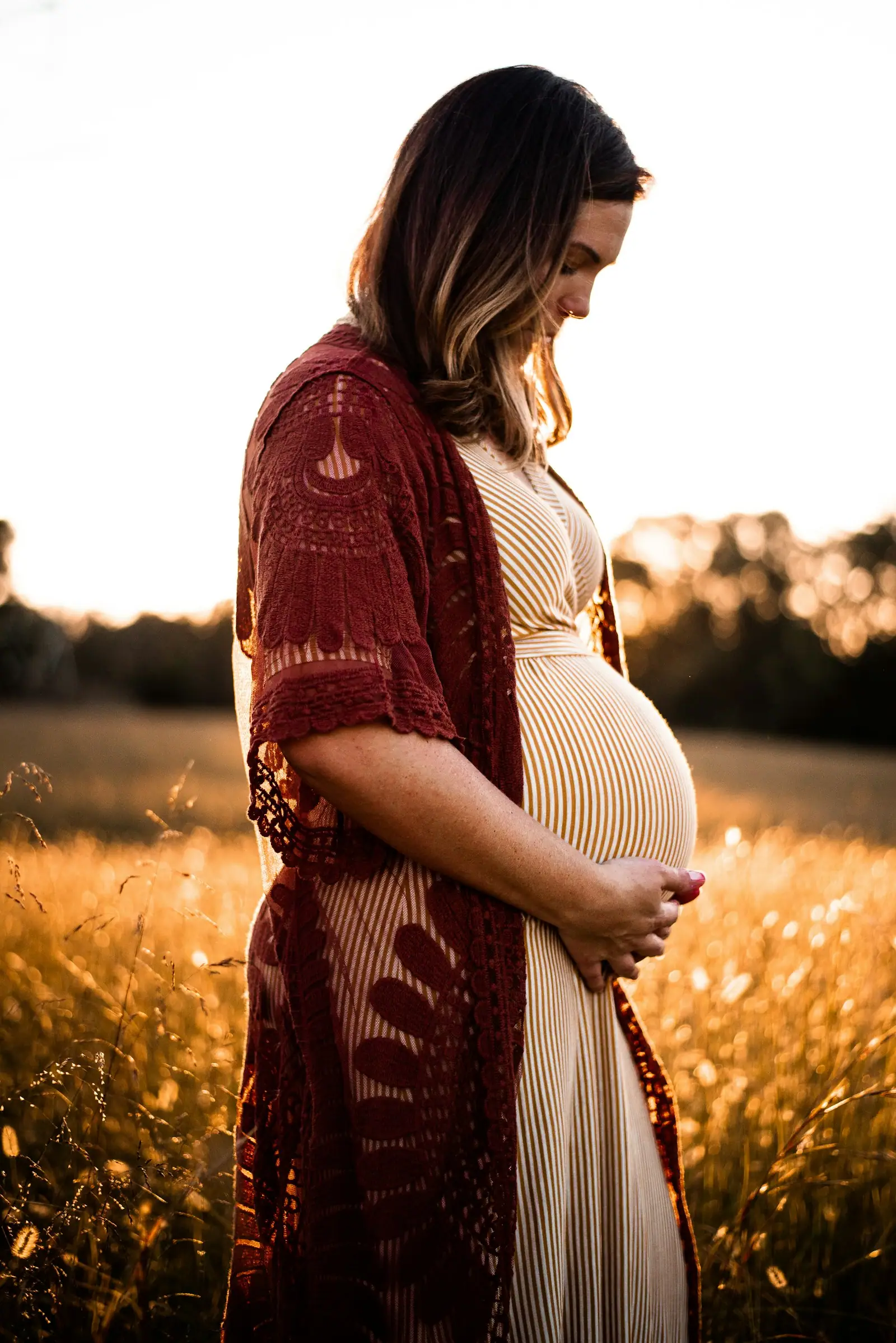 A pregnant woman in a striped dress and lace cardigan in a sunlit field