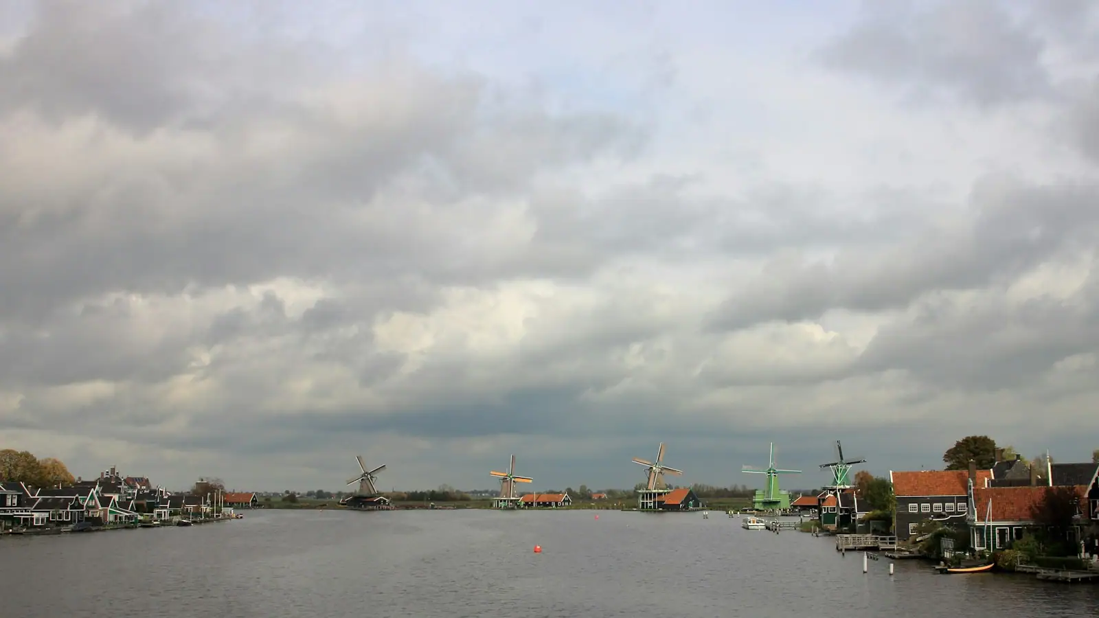 Windmills and houses line a river under cloudy skies