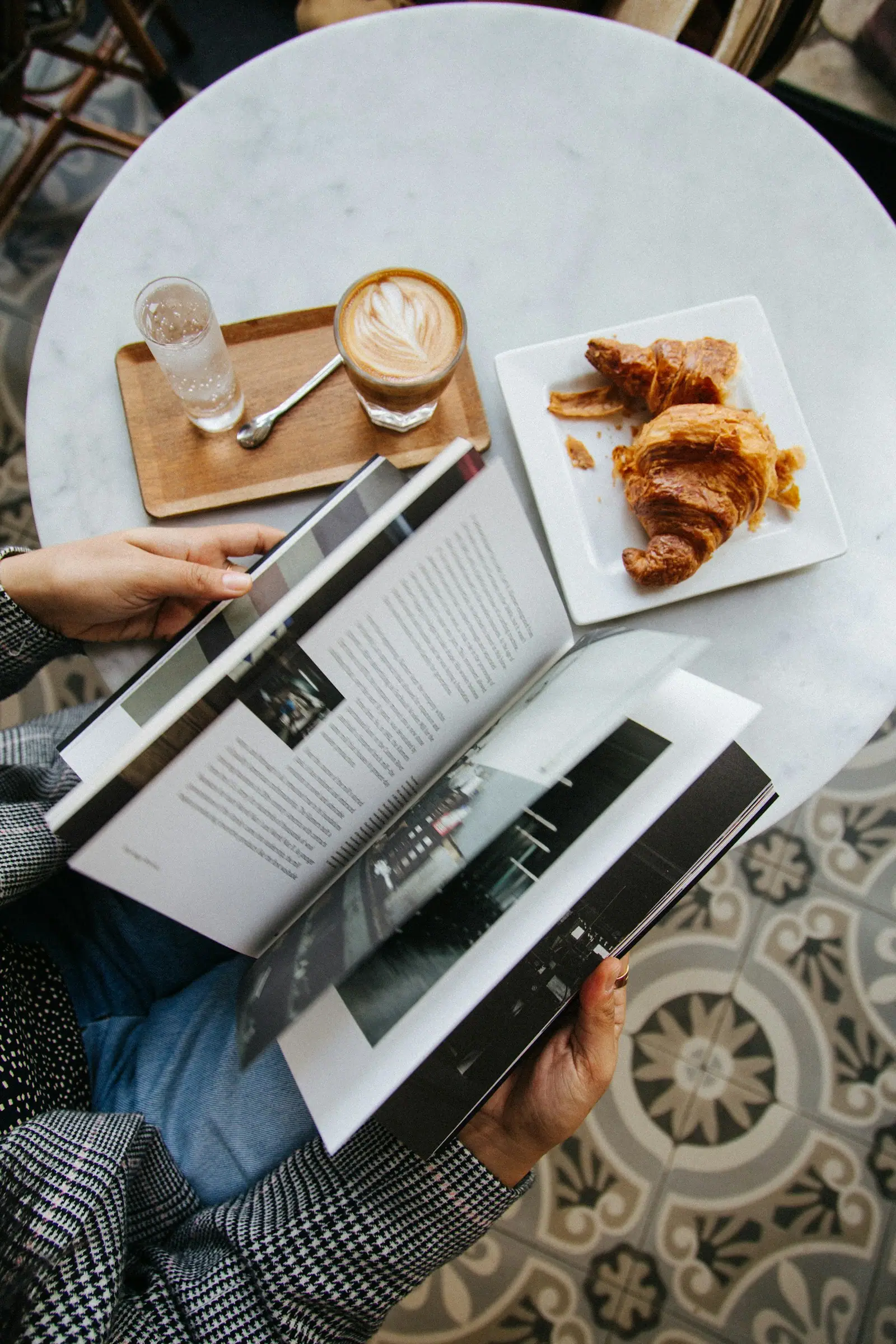 A person reading a book beside a coffee and croissants on a table